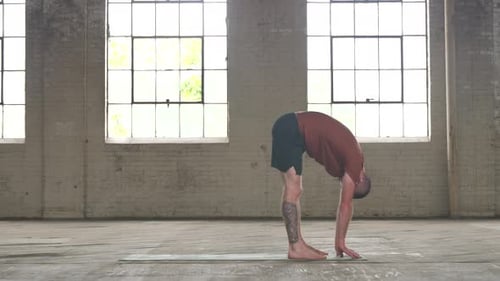 Mann in einer Industriehalle praktiziert Yoga, die Pose der Vorwärtsfalte, und greift nach seinen Zehen.