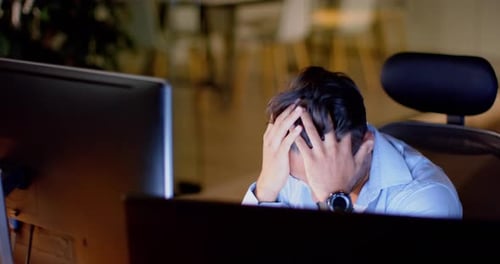 Tired biracial businessman at desk with computers in office at night, slow motion