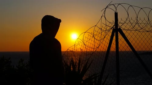 Silhouette of Young Adult Behind Barbed Wire Fence at Sunset