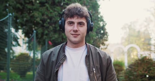 Happy Young Caucasian Man in Headphones and Jacket Walking the Street and Listening to Music
