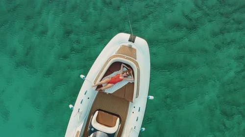 Vue aérienne d'une jeune fille blonde en maillot de bain rose prenant un bain de soleil sur un bateau à moteur entouré d'eau claire.
