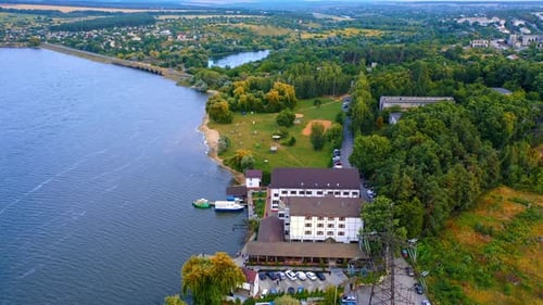 Green wooded bank of the river locating in the city. Hotel with the pier and few boats on the water.