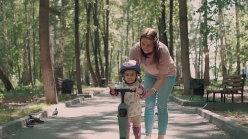 Mom Teaches Daughter to Ride a Scooter in the Park in Summer