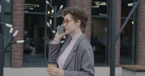 Side View of Business Lady Walking in Street Talking on Mobile Phone Holding to Go Coffee Cup