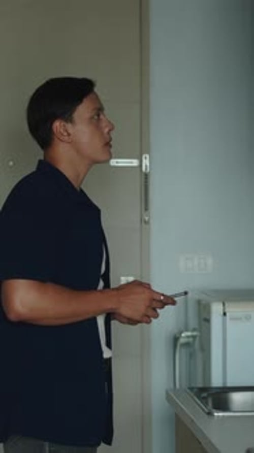Young Adult Man Standing In Modern Kitchen Looking At Smartphone Screen While Microwave Heats Fast