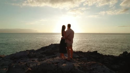 A couple stands on rocky terrain, embracing as they watch the sun set over the ocean, creating a sil