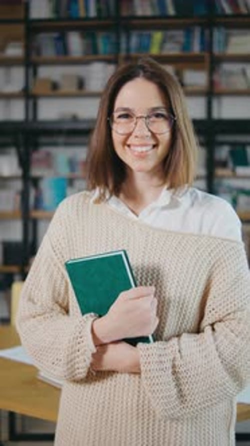 A Young Woman with Glasses in a Library Smiling Holding Book and Browsing Shelves Woman Holding Book