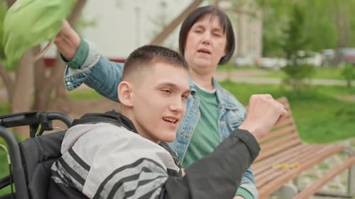 White Woman Flipping Cap On Teen In Wheelchair Cheerful Thumbs Up And Playful Interaction On Park