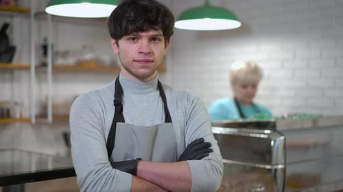 Portrait of Smiling Waiter Crossing Hands Looking at Camera As Barista Making Coffee at Background