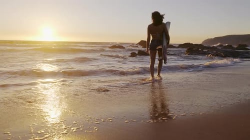 Woman Carries Surfboard on Beach at Sunset