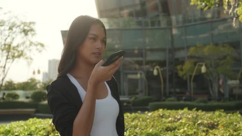 Portrait of Asian Lady with Loose Hair Sends Voice Message on Smartphone in Park