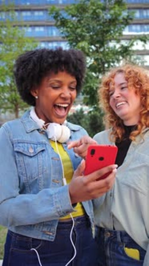 Two women friends look at phone and laugh outdoors