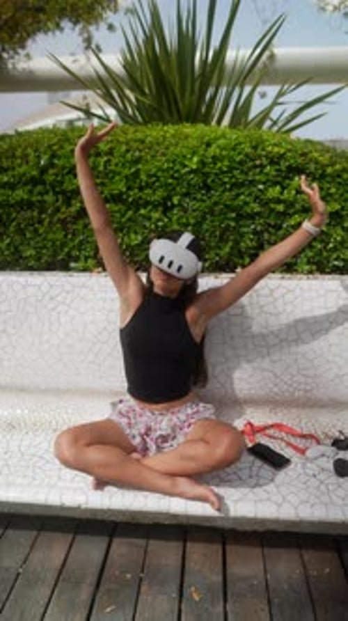 A Woman Uses VR Glasses to Do Yoga Outdoors on a White Bench in a Serene Garden