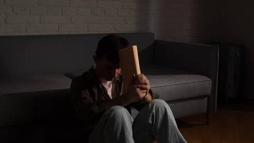 Man Sitting on the Wooden Floor in a Dimly Lit Room Holding a Blank Picture Frame Close to His Face