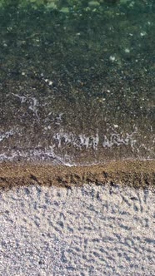 Vertikale Luftaufnahme des Strandes mit Wellen, die die Küste streicheln, Sonnenblendung, kleine Wellen auf der Wasseroberfläche