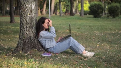Young Woman with a Laptop Sitting on the Grass in the Park on an Autumn Day