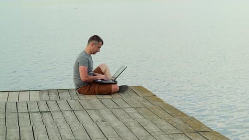 Handsome man in casual wear sitting on a wooden bridge over the blue water and works on a laptop