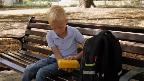 A Small Schoolboy with a Backpack Sits on a Park Bench and Eats a Sandwich