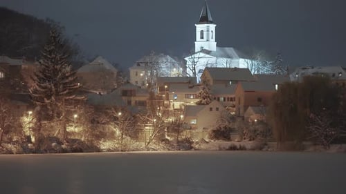 A peaceful Prague neighborhood at night features snow-covered trees and houses, with a church standi