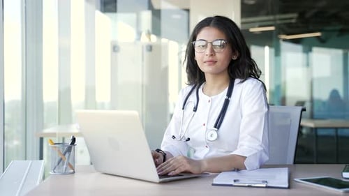 Portrait of young smiling female doctor working on laptop sitting at desk at workplace hospital