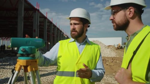 Two Surveyors Standing By Special Equipment and Looking at Total Station Men Dressed in Yellow Vests