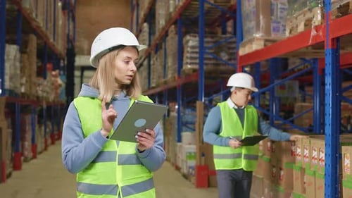 Lady Using Tablet While Man Noting on Clipboard at Warehouse