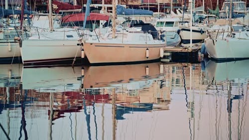 Sailboats and yachts docked in a calm marina, reflected in still water with pastel colored residenti