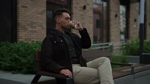 Happy Relaxed Young Man Sitting on City Bench Savoring Cup of Coffee Enjoying Pleasant Atmosphere of