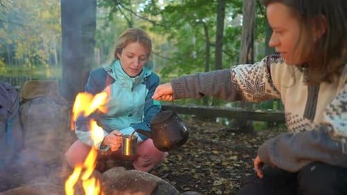Two Female Friends Make Coffee in a Kettle on an Open Fire in the Forest During a Hike