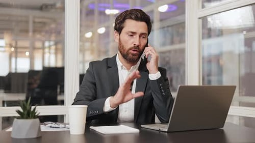 Businessman in Suit Talks on Cell Phone and Gestures to Laptop at Office Desk