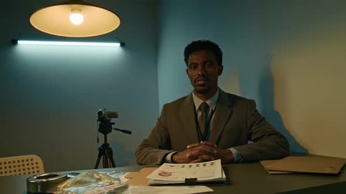 Low Key Portrait of Male Investigator Sitting at Table with Criminal Files