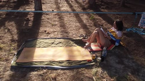 Young Woman Resting Reading Book In Camp In Wild Pine Forest At Sunset