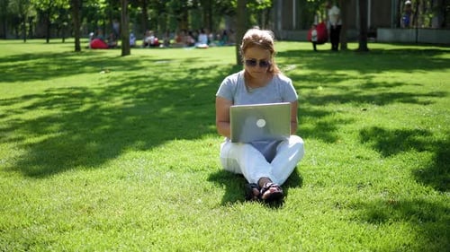 A Focused Woman in Sunglasses is Working with a Laptop in a Summer Park