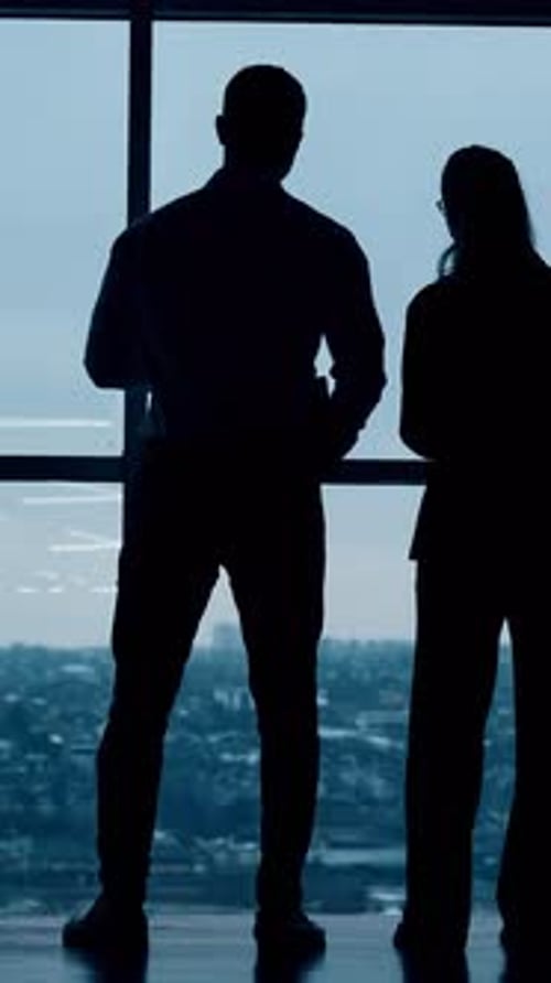 Dark silhouettes of a couple standing looking at the window. Colleagues in office look at cityscape.