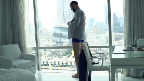 Man dressing in shirt in apartment overlooking city
