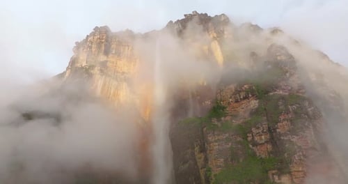 Massive Waterfall Descending From Sunlit Ancient Mountain Peaks Through Thick Clouds