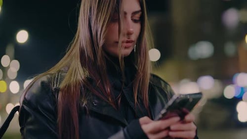 Young Woman Texts Message on Smartphone Standing on City Street Late in Evening