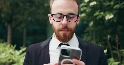 Close up. Cheerful man in glasses sitting on bench outdoors using typing smartphone. Confident beard