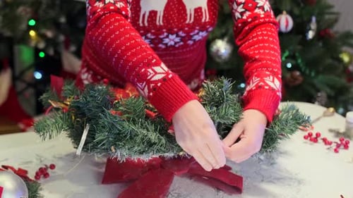 Woman hand in Christmas sweater with deer assembling spruce wreath decorating it with red bows
