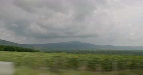 Side view of the mountains from the moving car on a cloudy day, with farmland in the foreground.