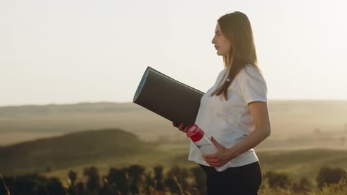 Schwangere Kaukasische Frau steht vor dem Hintergrund Sonnenuntergang hält Fitnessmatte und Flasche Wasser in