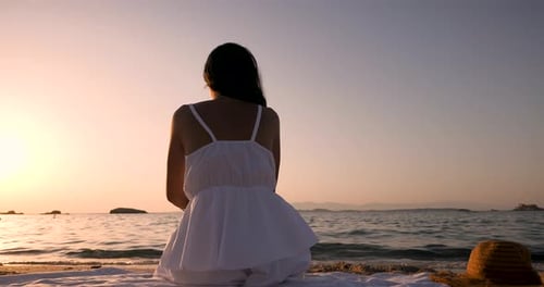 Back View shot of an young brunette woman tourist in white sundress is sitting on a sandy beach of