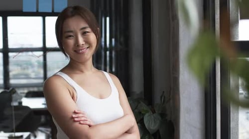 Asian woman standing by window in business office, arms crossed, looking confident, copy space