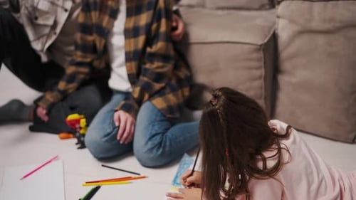 Close Up a Little Brunette Girl in Pink Clothes Draws Lying on the Floor Her Parents Look at Her a