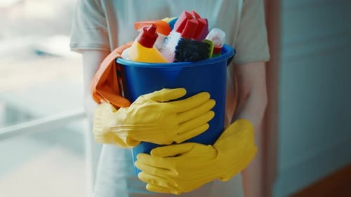 Cleaning Supplies in a Bucket Held By a Person Wearing Yellow Gloves Indoors During Daylight Hours