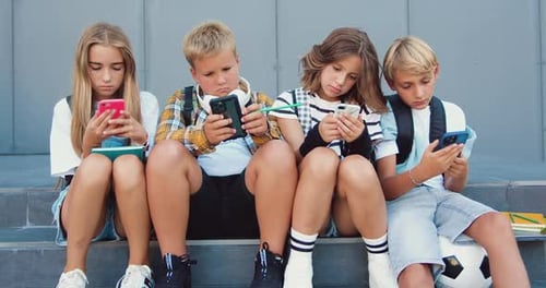 Group of teenage sad friends holding phones and browsing sitting on stairs near school building