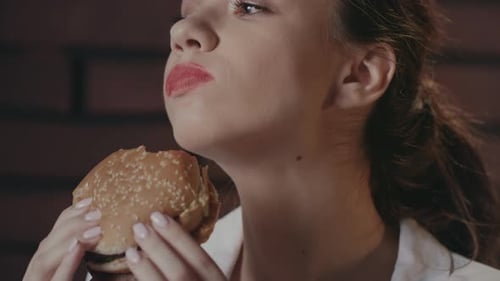 Hungry Woman Eating Burger On Brick Wall Background In Fast Food Cafe. Young Woman Eating Meat Bu...