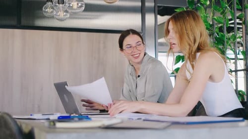 Two Professional Women Engage in a Collaborative Discussion at a Modern Office Table Surrounded By