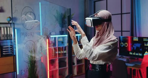 Attractive woman in hijab working on imaginary screen in virtual reality headset in evening office.