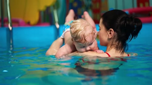 Rear view of a woman holding a baby in the swimming pool. Coach is teaching an infant to swim.